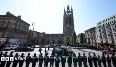 A funeral cortege arrives at Newcastle Cathedral as rows of police officers stand in formation, with mourners gathered outside the church during a ceremonial service. It is a sunny day. Attendees stand on the edge of the square in front of the cathedral, with clergy waiting for the cortege in the centre of the plaza.