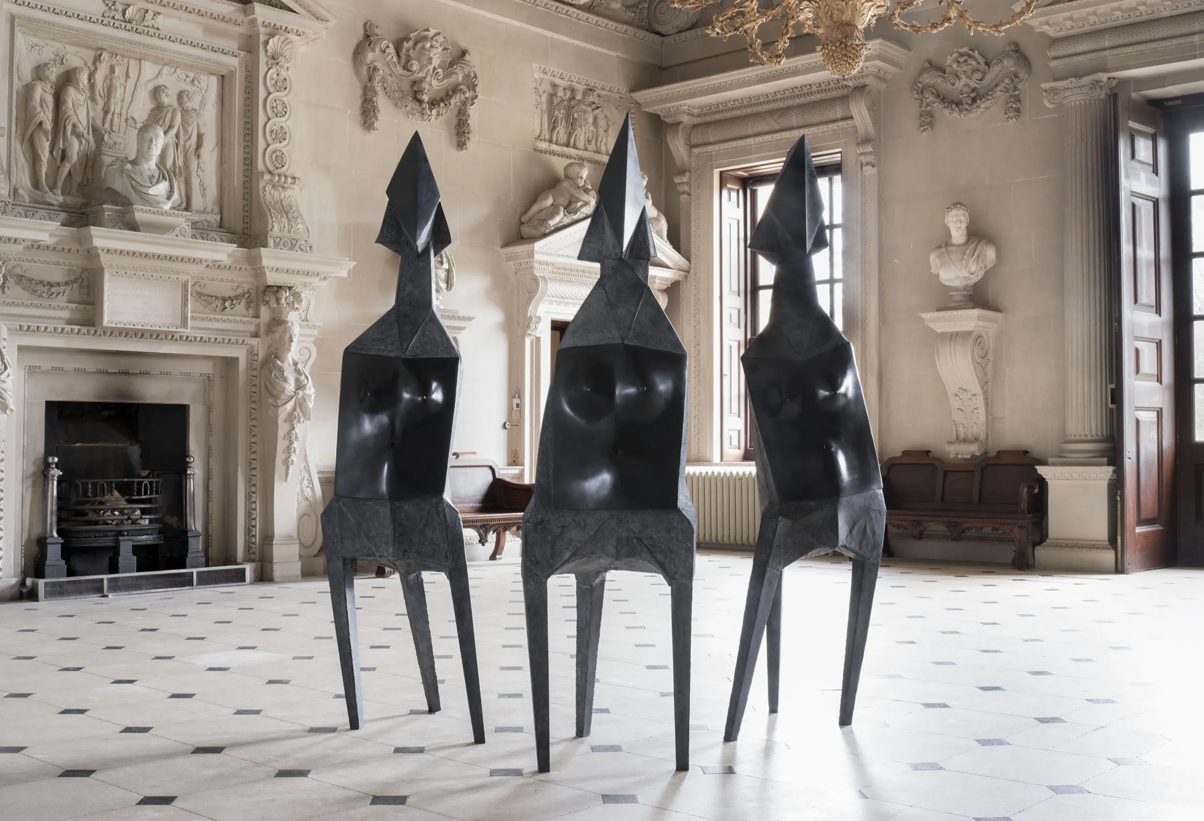 Three black abstract sculptures by Lynn Chadwick titled "Elektras" stand in a grand room with ornate white walls and a tiled floor.