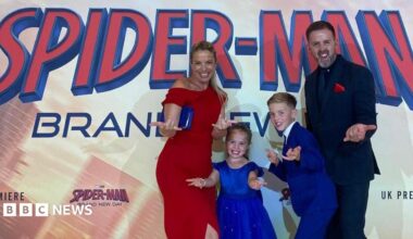 A blond woman in a red dress, man in a black suit, and two children in royal blue do the spider man pose, extending their hands to the camera, in front of the premiere poster.