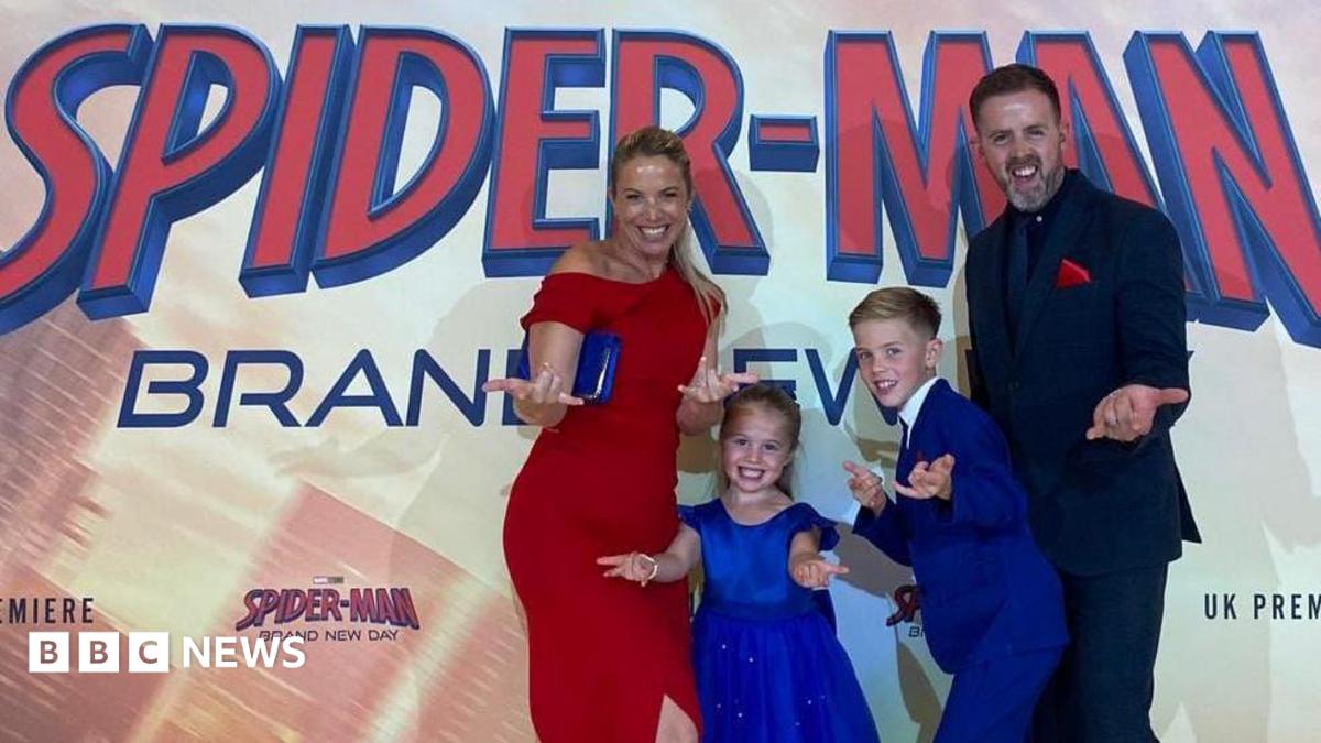A blond woman in a red dress, man in a black suit, and two children in royal blue do the spider man pose, extending their hands to the camera, in front of the premiere poster.