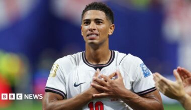 Image shows Jude Bellingham, a man with short afro hair, clutching his chest, on the pitch while playing for England. He is smiling. He wears a white England shirt with a red number 10.