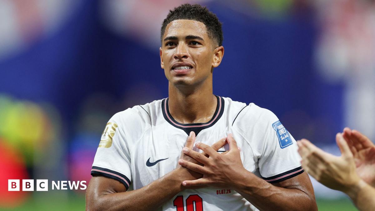 Image shows Jude Bellingham, a man with short afro hair, clutching his chest, on the pitch while playing for England. He is smiling. He wears a white England shirt with a red number 10.