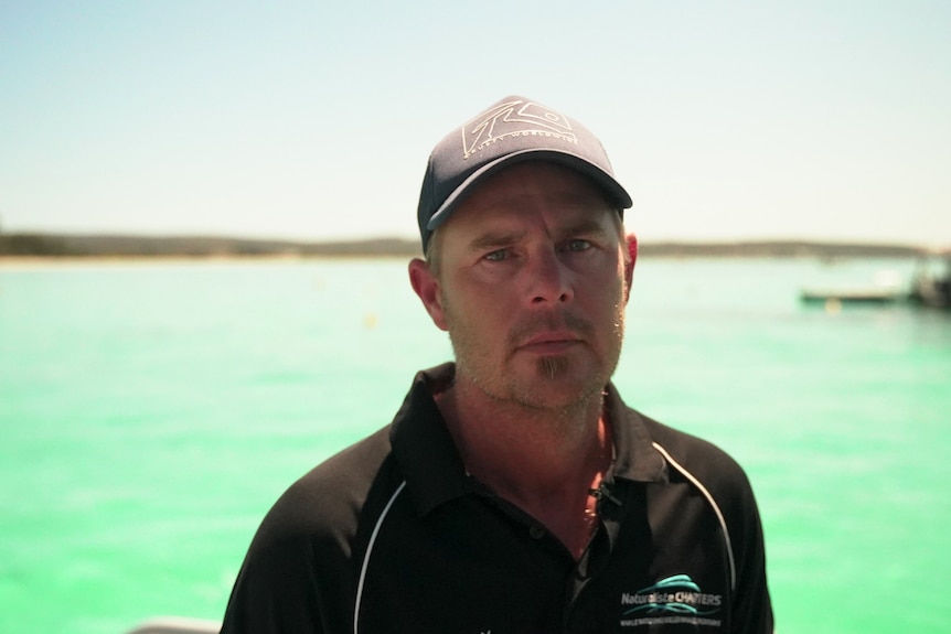 A man with a goatee and a cap in a black polo short standing in front of green beach water.