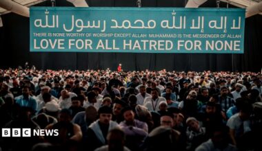A crowd of people sat in a building under a large banner which says "There is none worthy of worship except Allah Muhammad is the messenger of Allah" "Love for all Hatred for none"