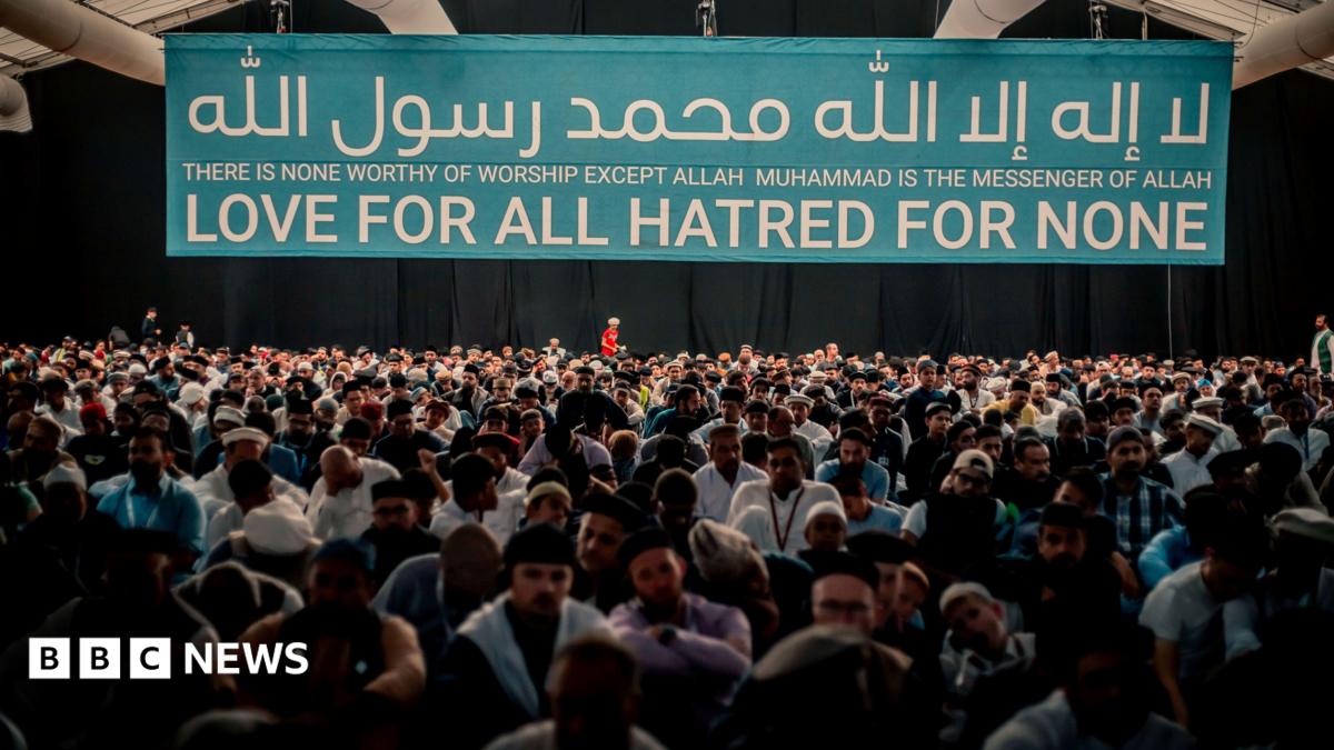 A crowd of people sat in a building under a large banner which says "There is none worthy of worship except Allah Muhammad is the messenger of Allah" "Love for all Hatred for none"