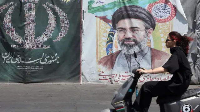 A woman on a scooter driving past a banner of Mojtaba Khamenei