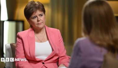 Nicola Sturgeon wearing a pink suit and white vest, with a quizzical expression. She is sitting on a chair facing another woman who has blonde hair and is wearing purple.