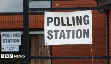 A red-brick building with a paper sign tied to black railings. The sign reads: 'POLLING STATION'.