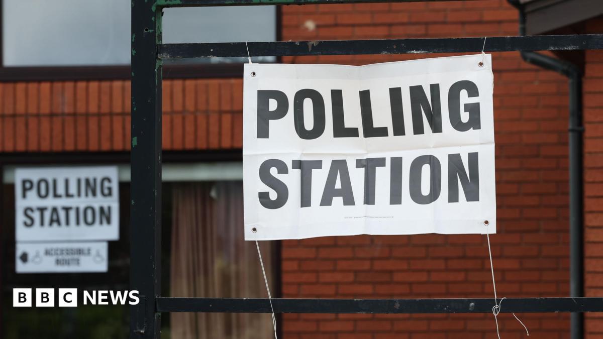 A red-brick building with a paper sign tied to black railings. The sign reads: 'POLLING STATION'.