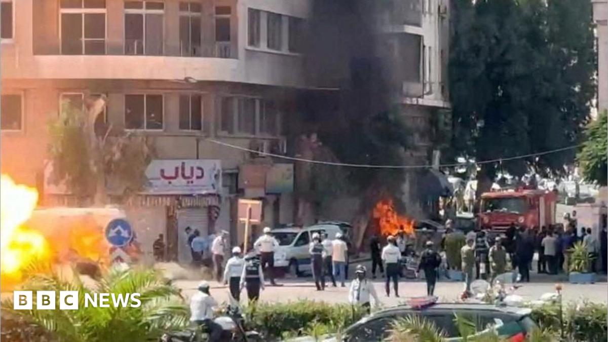 A wide shot of a street with lots of emergency workers. Flames and smoke rise from an alleged explosion site