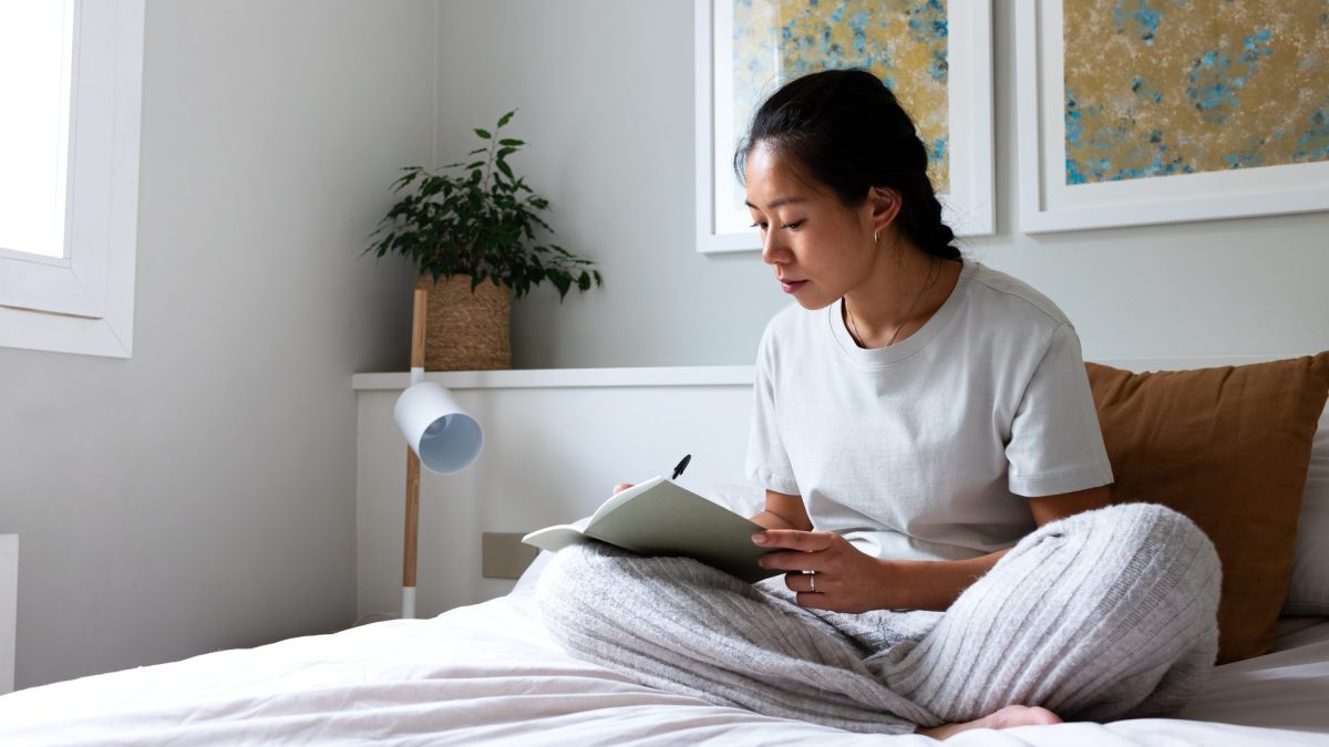 Pensive young Asian woman sitting on bed writing on journal in cozy bedroom. Copy space. - stock photo