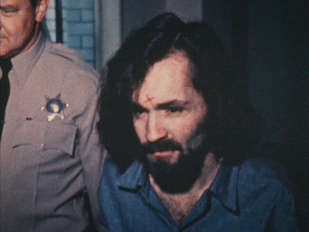 A man with shoulder length dark hair and a beard in a blue prison jumpsuit being walked with a sherriff.