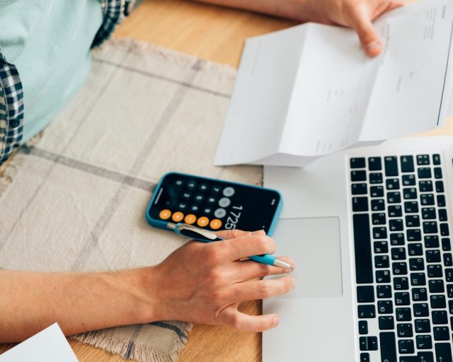 Unrecognizable person counting expenses on calculator in mobile phone app, checking her energy bills at home. Budget management, price increases, inflation, rise in price of products and utilities. View from above, managing home finances - stock photo