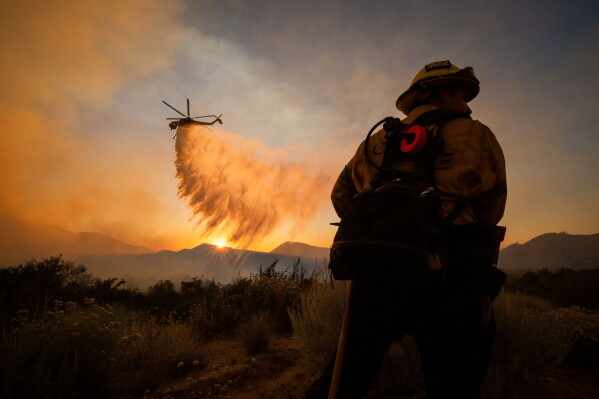 A firefighter watches as a helicopter drops water on the Summit Fire while battling wildfires in Llano, Calif., July 10, 2026. (AP Photo/Ethan Swope, File)
