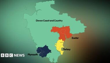 A man wearing a short sleeved blue shirt is standing on a pavement outdoors. Buildings are visible in the blurred background behind him.