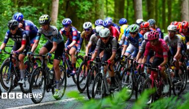 A tightly packed group of road cyclists on a road surrounded by woodland.