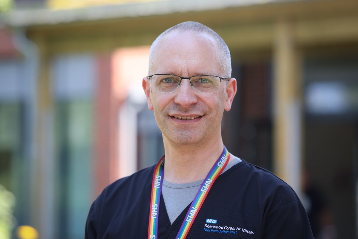 Middle-aged man with nearly-shaved head and glasses, wearing nurses scrubs and rainbow NHS lanyard, smiling at camera in front of colourful blurred outdoor background