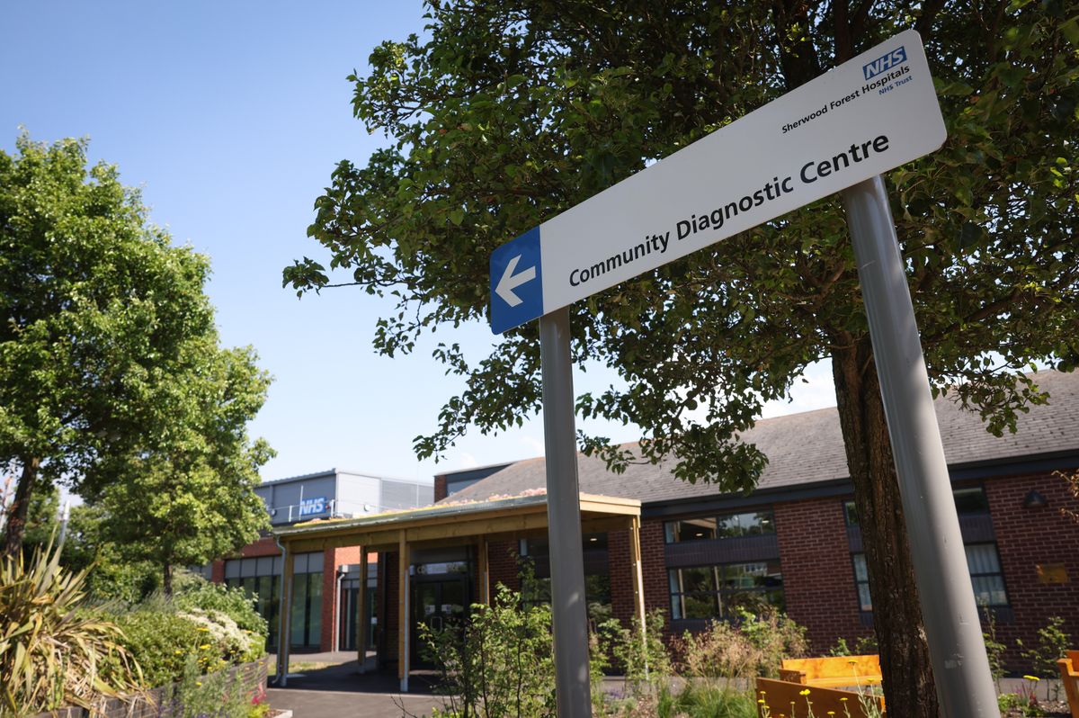 White NHS sign with white arrow on blue background in corner, outside modern brick building with large NHS sign on facade in background, with trees, bushes and greenery visible in garden on way to entrance in foreground