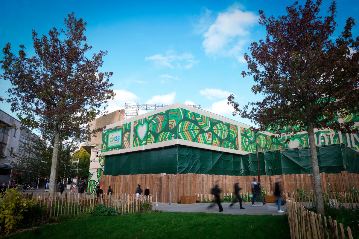 Green tarpaulin/sheeting and fencing covering floors of old building, with crane/scaffolding visible above in background, and green public space with fencing and trees visible in foreground, with people walking past on footpaths through, and quiet city centre pedestrian street visible in background to left, pictured on sunny day with blue sky and clouds