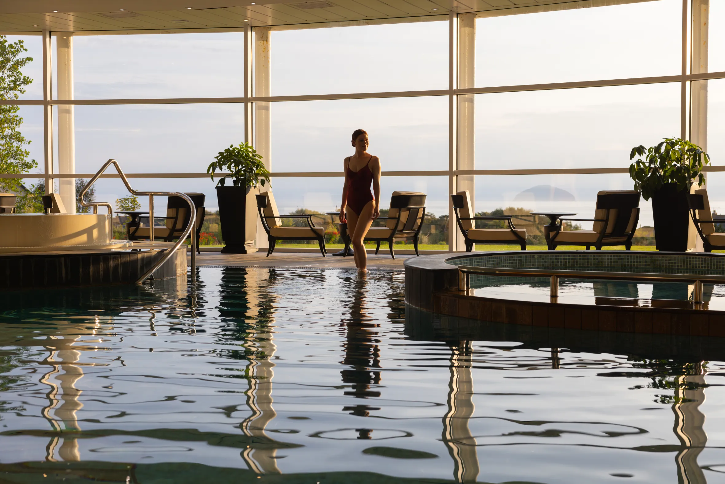 A woman in a red swimsuit stands in a shallow pool in an indoor spa with large windows overlooking a green landscape.