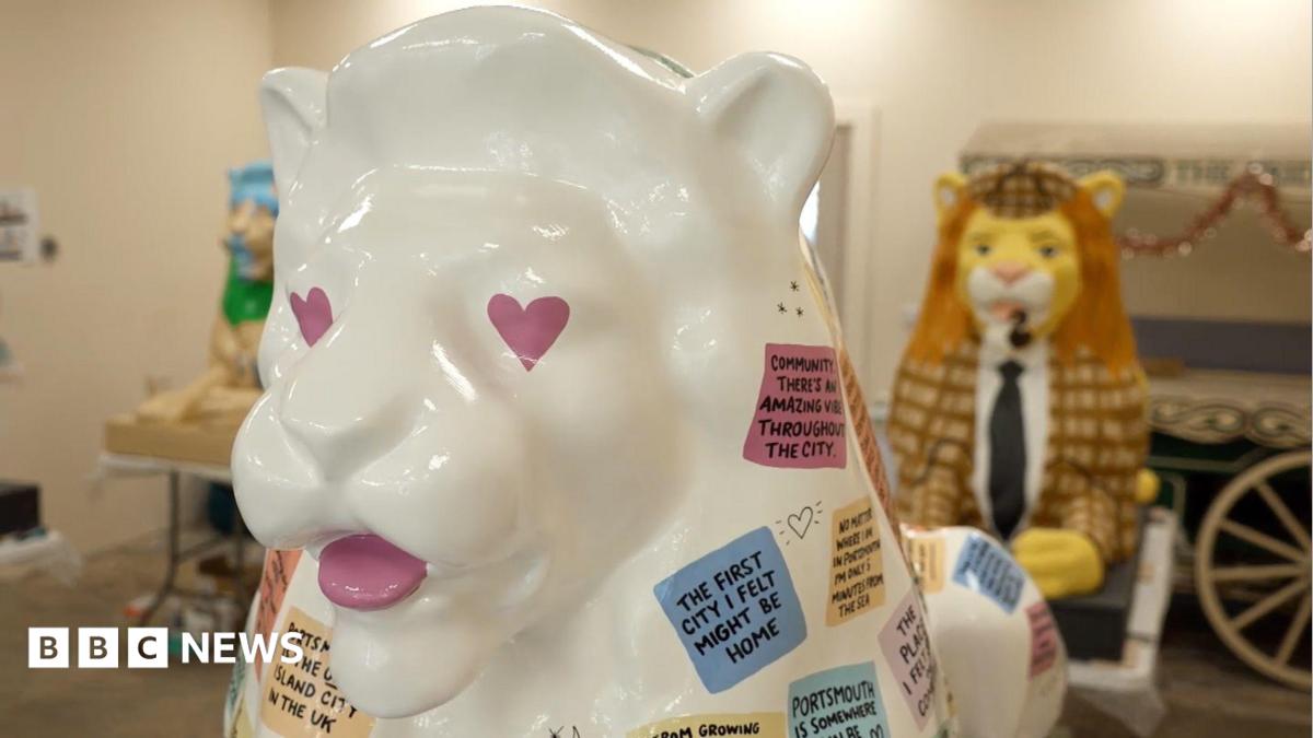 The image shows three lion sculptures. Two in the background are slightly blurred and the one if the foreground is in focus. The one in the front is white and painted with pink heart eyes. It has a number of different coloured squares which each say different messages about Portsmouth. The one in the background is painted in to look like it is wearing a suit. It has a brown jacket on and a black tie. It has a ginger mane and a brown hat on. The third lion is inspired by history of libraries. It is a brown lion, which has green and grey on it.
