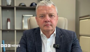 Darragh Cullen, a man with short, white hair, looks tot he camera with a serious expression.  He is wearing a navy checked suit jacket and a white shirt.  He is sitting in an office.