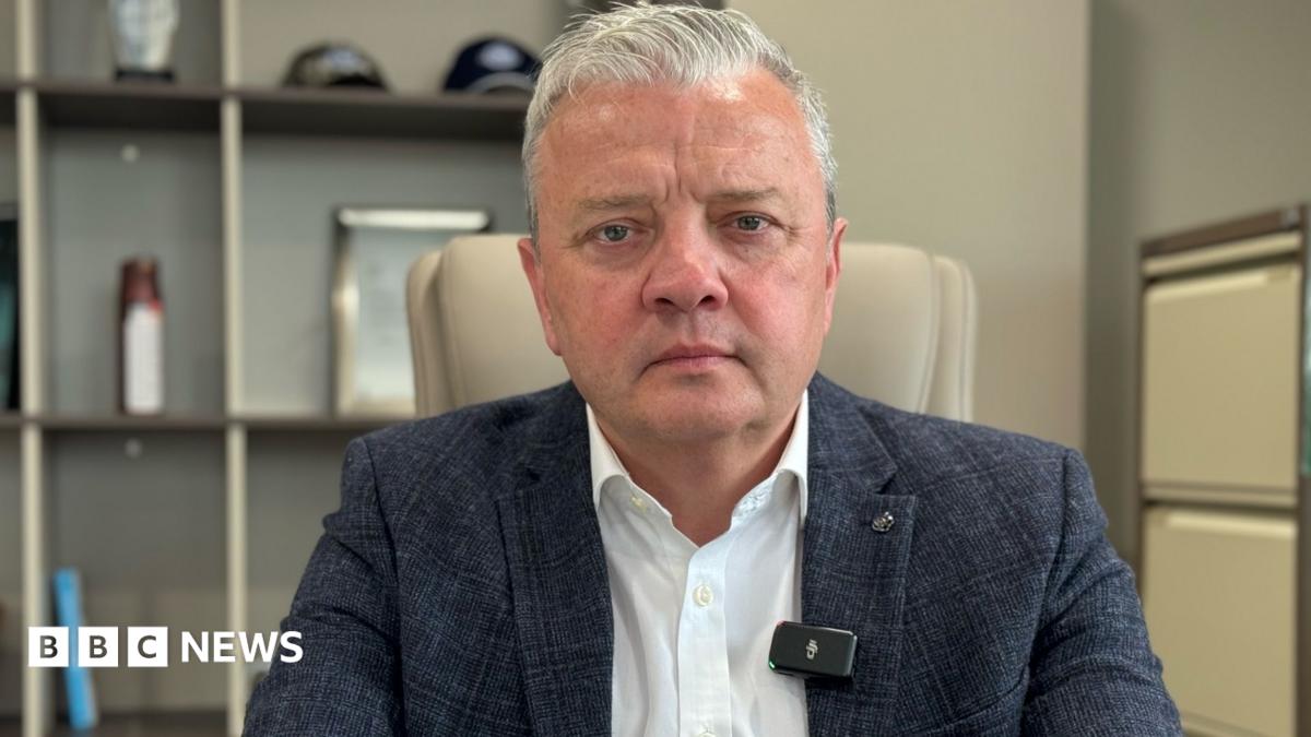 Darragh Cullen, a man with short, white hair, looks tot he camera with a serious expression.  He is wearing a navy checked suit jacket and a white shirt.  He is sitting in an office.