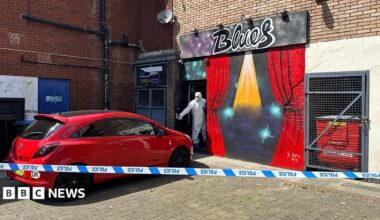 Police tape cordon around a red Corsa car parked outside a bar with the sign in graffiti saying 'Blues'. A CSI police officer wearing white personal protective equipment (PPE) is standing in the entrance holding the front door open by its handle.