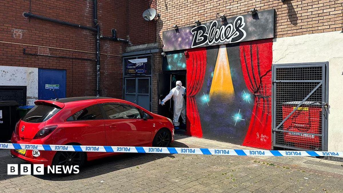 Police tape cordon around a red Corsa car parked outside a bar with the sign in graffiti saying 'Blues'. A CSI police officer wearing white personal protective equipment (PPE) is standing in the entrance holding the front door open by its handle.