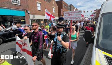 A demonstration waving England flags walking down a high street lined by brick buildings. One placard is visible, which reads "Illegal immigrants are not welcome. Stop the boats."