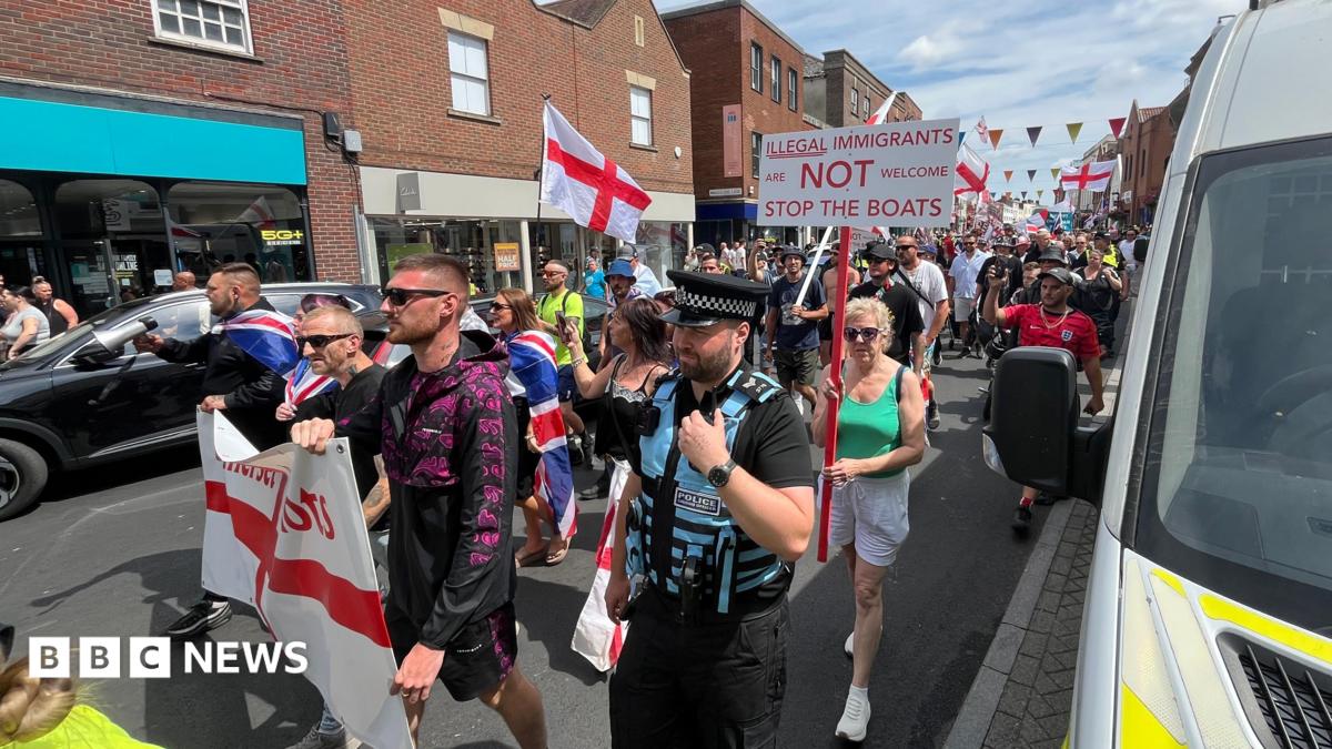 A demonstration waving England flags walking down a high street lined by brick buildings. One placard is visible, which reads "Illegal immigrants are not welcome. Stop the boats."