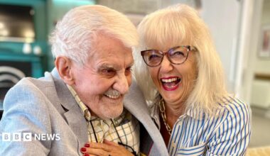 Clifford Craske and Alice Craske are sitting next to each other. Clifford, on the left, has short white hair and is wearing a checked shirt and a grey blazer. Alice, on the right, has shoulder-length blonde hair and a fringe, and is wearing a blue and white striped shirt and a gold necklace. Both are smiling.