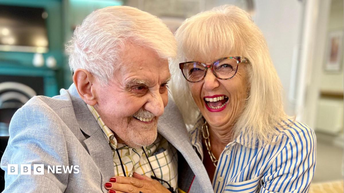 Clifford Craske and Alice Craske are sitting next to each other. Clifford, on the left, has short white hair and is wearing a checked shirt and a grey blazer. Alice, on the right, has shoulder-length blonde hair and a fringe, and is wearing a blue and white striped shirt and a gold necklace. Both are smiling.