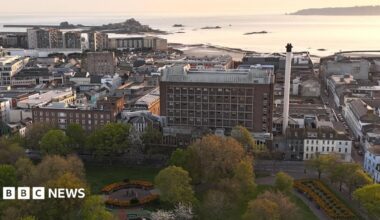 This is an elevated view of St Helier showing a large public park in the foreground, with trees and flower beds. The General Hospital is in view behind and other buildings and the sea are beyond. The coastline, harbour and sea stretch into the distance under warm evening light, with Elizabeth Castle visible on a tidal island offshore.