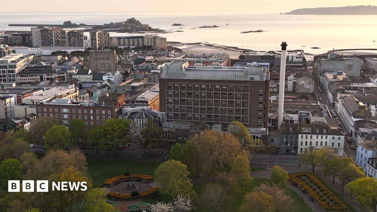 This is an elevated view of St Helier showing a large public park in the foreground, with trees and flower beds. The General Hospital is in view behind and other buildings and the sea are beyond. The coastline, harbour and sea stretch into the distance under warm evening light, with Elizabeth Castle visible on a tidal island offshore.