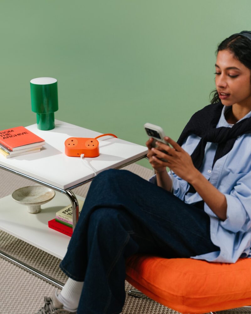 A woman sits in an orange chair using her phone next to a white table with books, a green lamp, an orange DailyObjects power strip, and a bowl on it.
