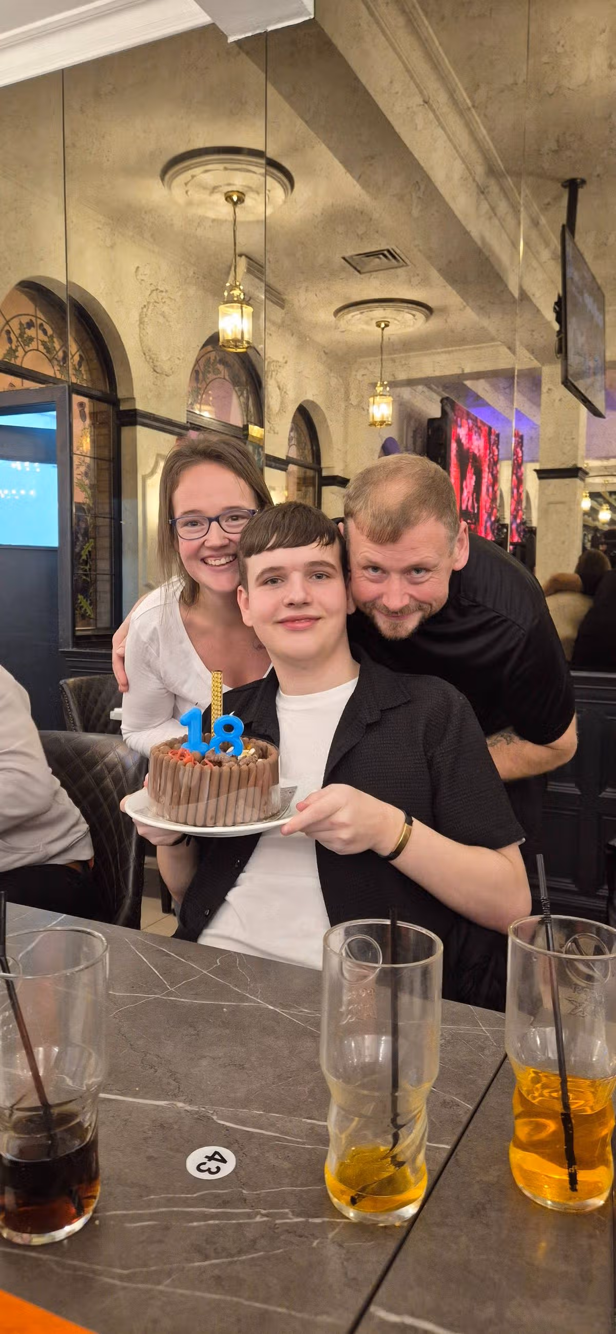 A family of three individuals, including a young man celebrating his 18th birthday, is gathered around a cake with a candle inscribed "18". They are smiling and posing for a photograph in a casual indoor setting.