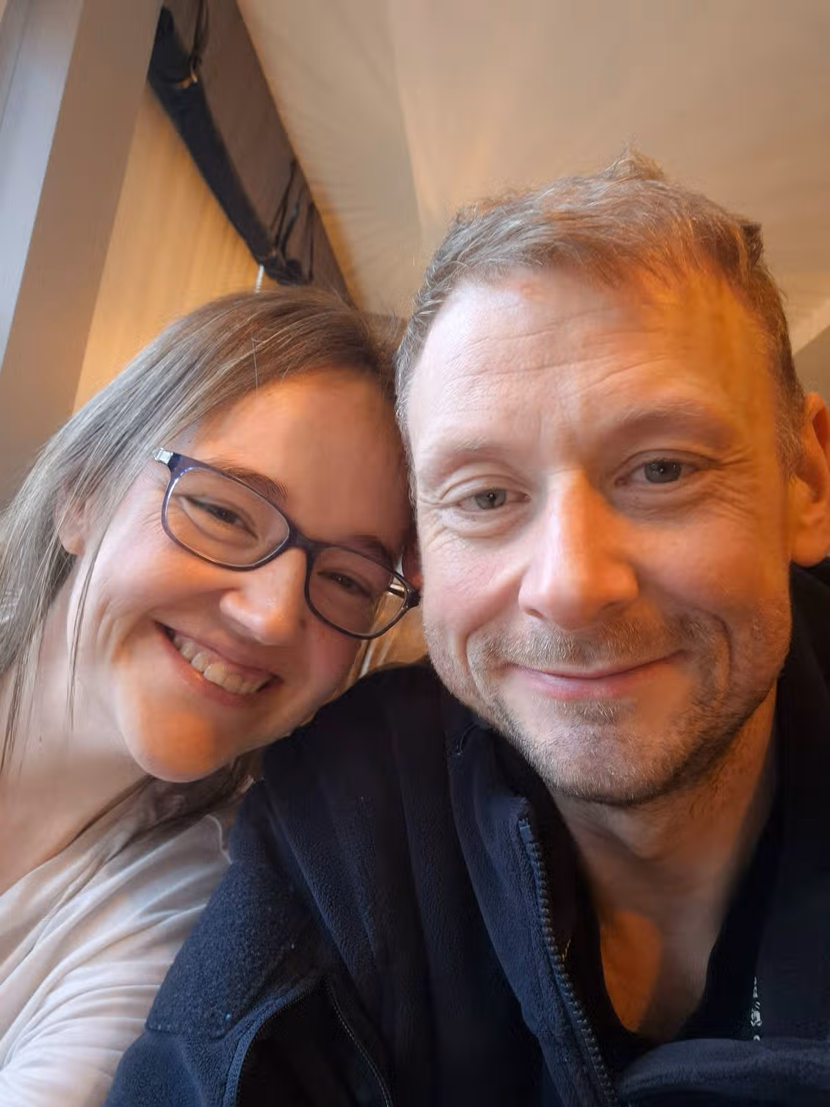 An image depicts two individuals, one on the left with long hair and glasses, and the other on the right, both wearing casual attire. They are posing closely together, with expressions of joy and contentment evident on their faces, suggesting a moment of shared happiness.