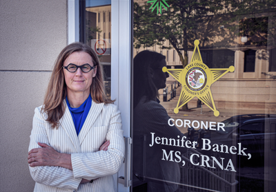 A picture of a woman with light brown hair wearing glasses and a white striped suit jacket. She's standing with arms crossed in front of a door that reads "Coroner Jennifer Banek, MS, CRNA."