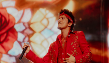 Bruno Mars holds a microphone and performs at Wembley stadium wearing a red suit with gold flower detail. He also wears a red headband.
