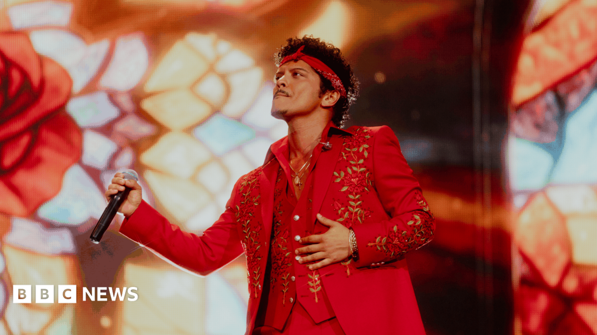 Bruno Mars holds a microphone and performs at Wembley stadium wearing a red suit with gold flower detail. He also wears a red headband.