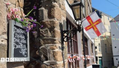 A chalkboard sign reading “Bonjour, Bienvenue dans notre Boutique” mounted on a stone wall beneath pink and purple flowers. A Guernsey flag hangs from a black lantern bracket, with bunting visible along the street in the background
