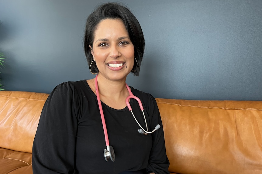 A woman in a black shirt and black hair and a stethoscope around her neck smiles.