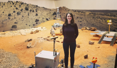 A woman wearing a brown jumper and black trousers stands on a simulated surface of Mars with orange sand and stones, with prototypes at her feet
