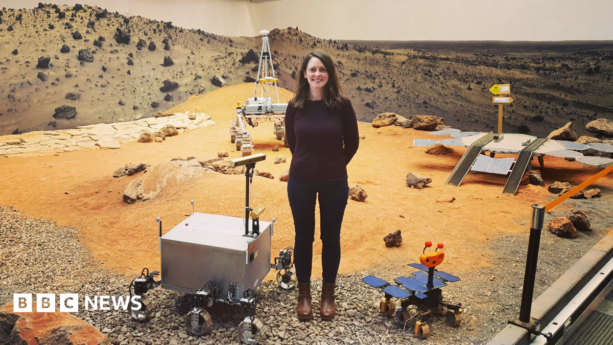 A woman wearing a brown jumper and black trousers stands on a simulated surface of Mars with orange sand and stones, with prototypes at her feet