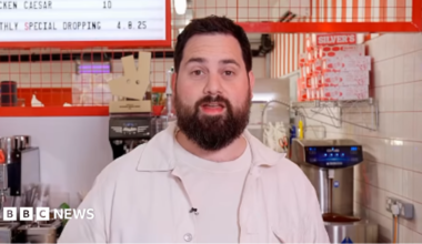 A picture of Rich Myers standing in his shop, behind a counter. There is a coffee machine and menu in the background.