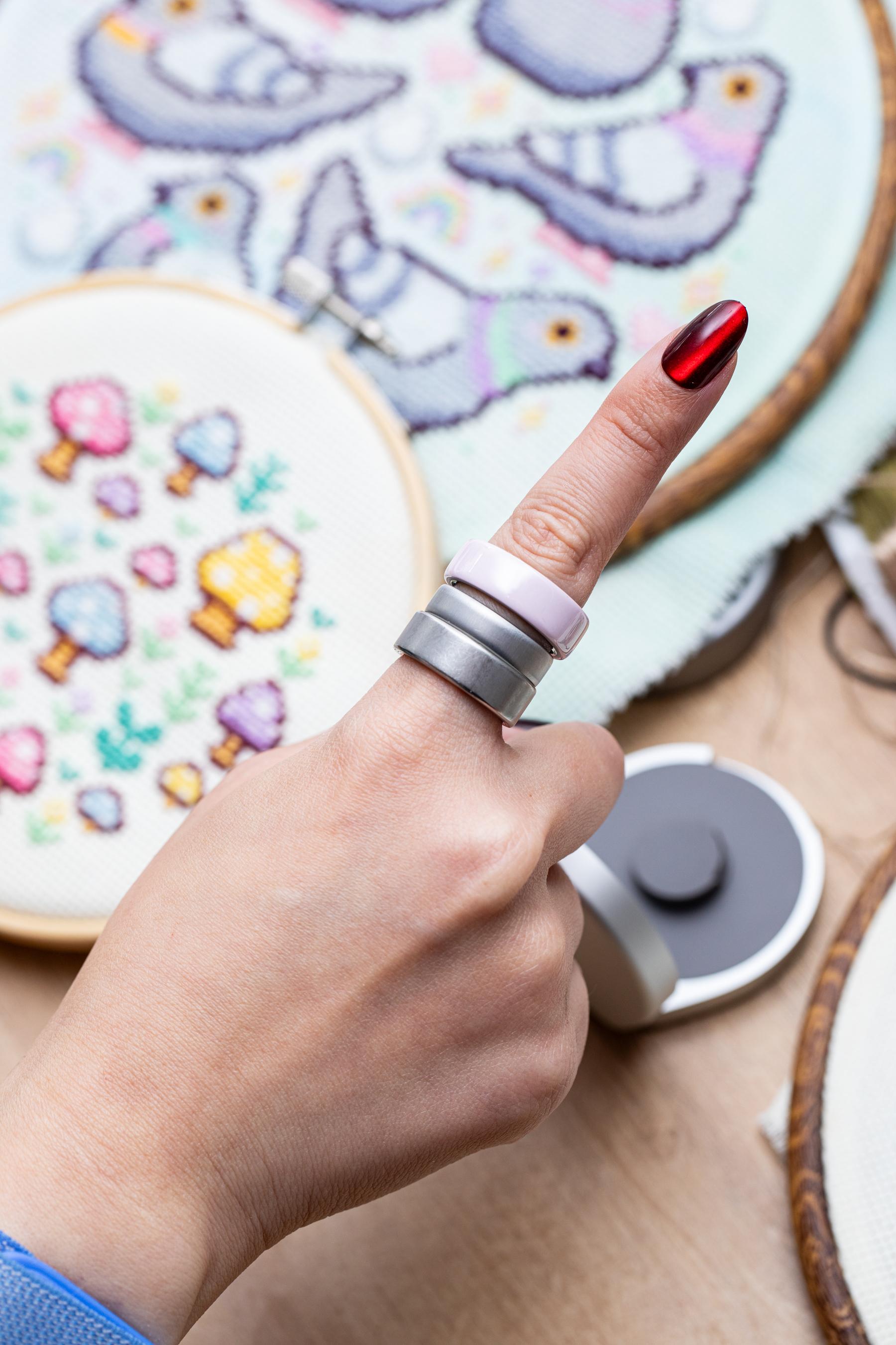 Someone wearing two Oura Ring 4s with an Oura Ring 5 in beween with cross stitch projects in the background.