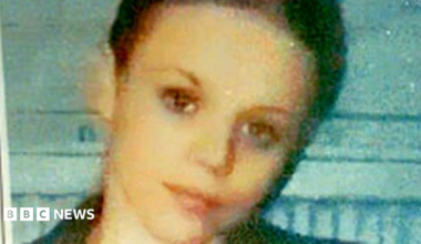 Donna Keogh with cropped dark hair and dark eyes. She is smiling with her head tilted to one side and standing in front of a light blue wall. The photo is very grainy.