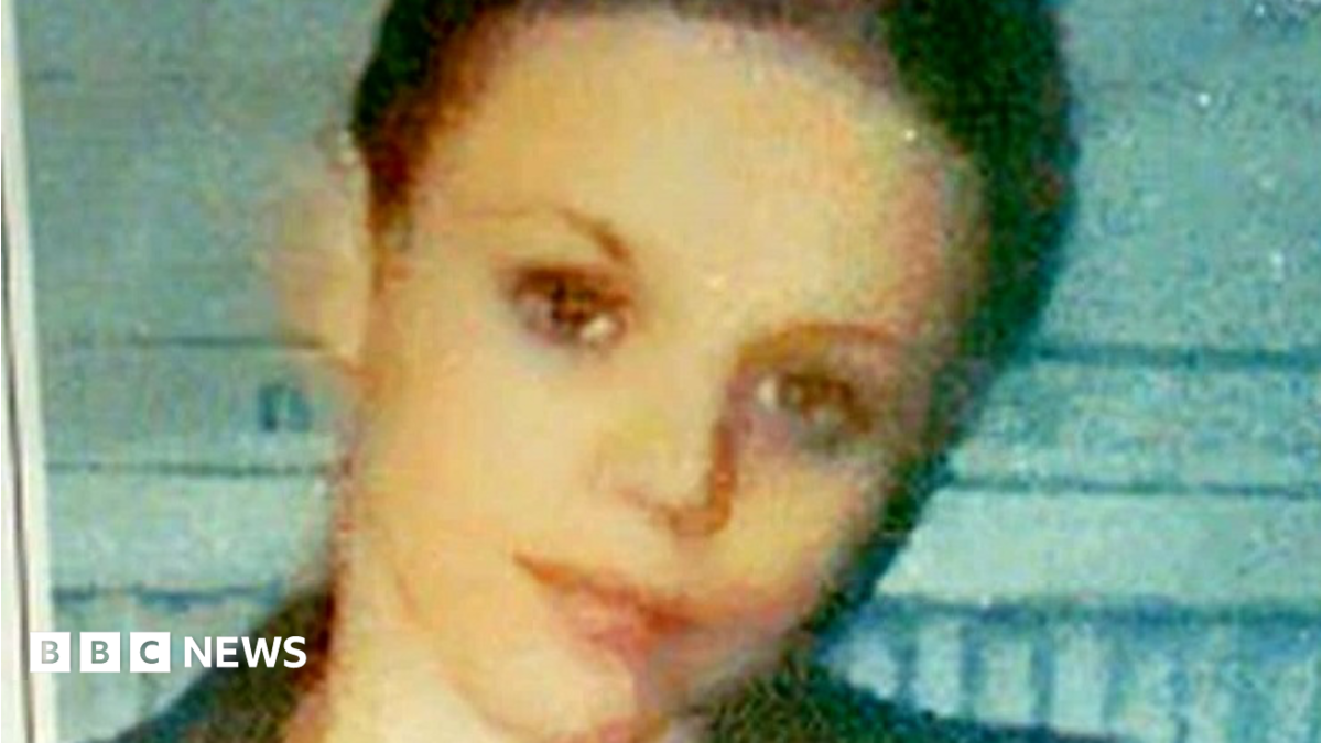Donna Keogh with cropped dark hair and dark eyes. She is smiling with her head tilted to one side and standing in front of a light blue wall. The photo is very grainy.