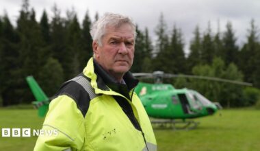 Nethy Bridge community council leader Sandy McCook wearing a high-visibility yellow jacket stands in a grassy clearing with a green helicopter parked behind them. Dense conifer forest forms the background under an overcast sky.
